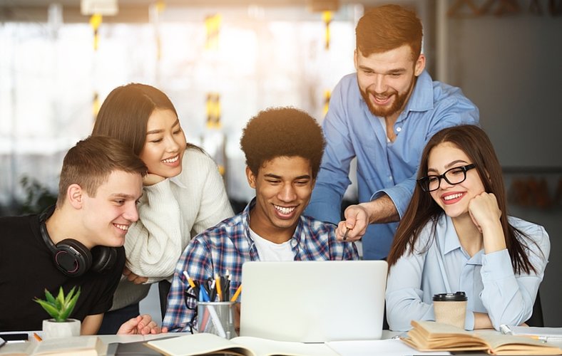 Fünf Student*innen unterschiedlichen Aussehens sitzen bzw. stehen an einem Bürotisch und blicken auf einen Laptop. Sie lachen fröhlich. Auf dem Tisch stehen ein Kaktus und ein Coffee-to-Go-Becher, außerdem liegen Bücher darauf.