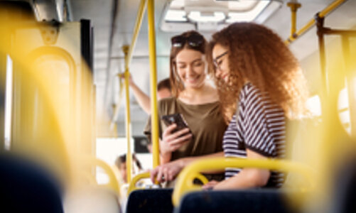 Frauen im Bus Zwei junge Frauen stehen in einem Bus und schauen lächelnd auf ein Handy. Sie tragen kurze T-Shirt, eine der beiden hat eine Sonnenbrille im Haar. Die Haltestangen des Busses bringen gelbe Farbkleckse ins Bild.