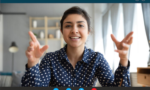 Im Web-Meeting Eine junge Frau in einer dunkelblauen Bluse mit weißen Punkten sitzt vor einem Bildschirm und gestikuliert mit den Armen. Sie scheint Teilnehmerin eines Web-Meetings zu sein. Im Hintergrund ist ein minimalistisch eingerichtetes Zimmer erkennbar.