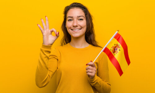 Spanien Ein junges Mädchen mit langen Haaren steht in einem gelben Pullover vor einer gelben Wand, ihre Fingernägel sind rot lackiert. In ihrer linken Hand hält sie die gelb-rote spanische Flagge, mit der rechten Hand signalisiert sie das "Perfekt"-Zeichen (Daumen und Zeigefinger sind zu einem Kreis geformt, die anderen Finger nach oben gereckt).